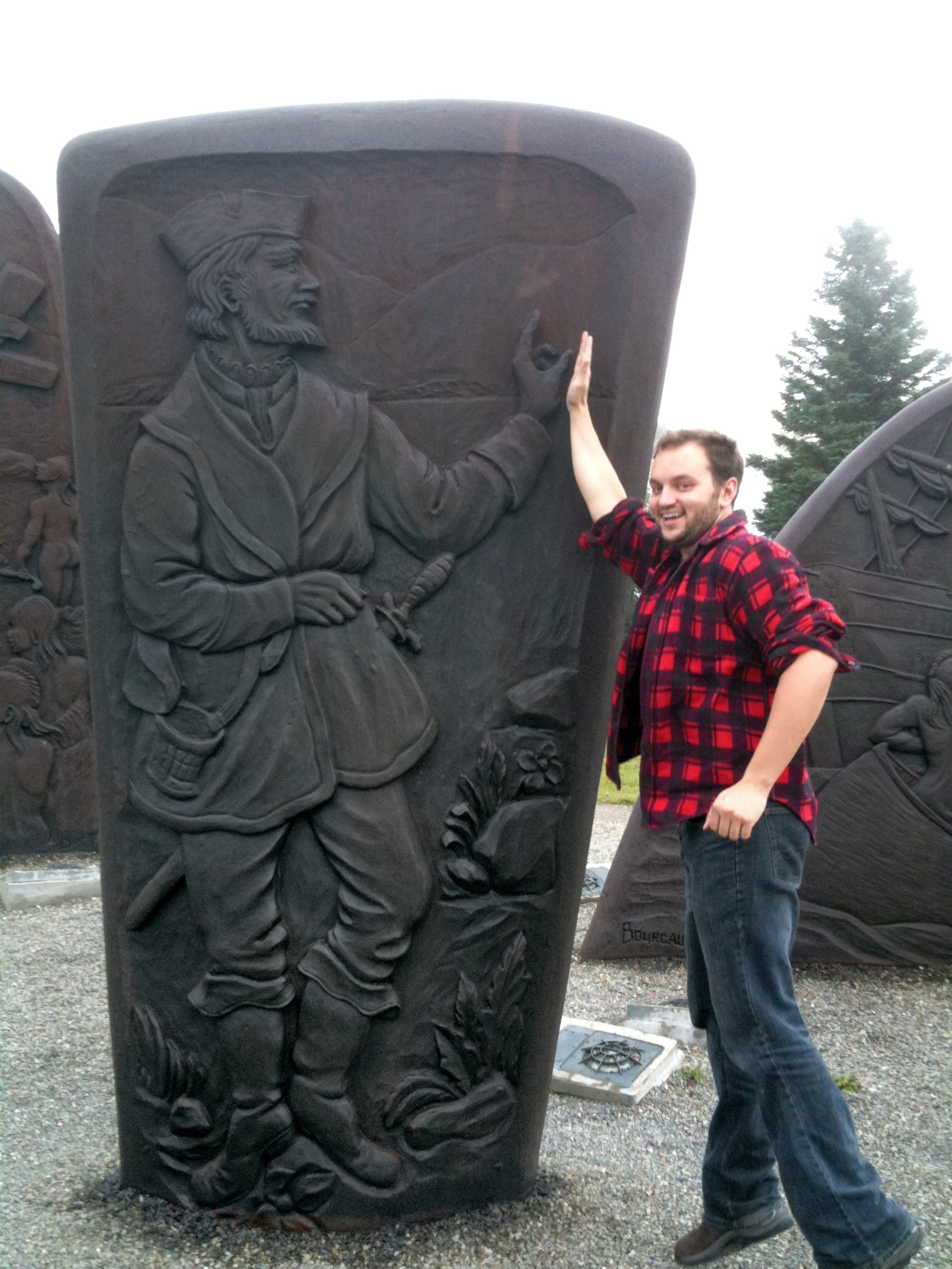 Simon Devost wearing a lumberjack shirt high-fiving a sculpture of Samuel de Champlain near Gaspé