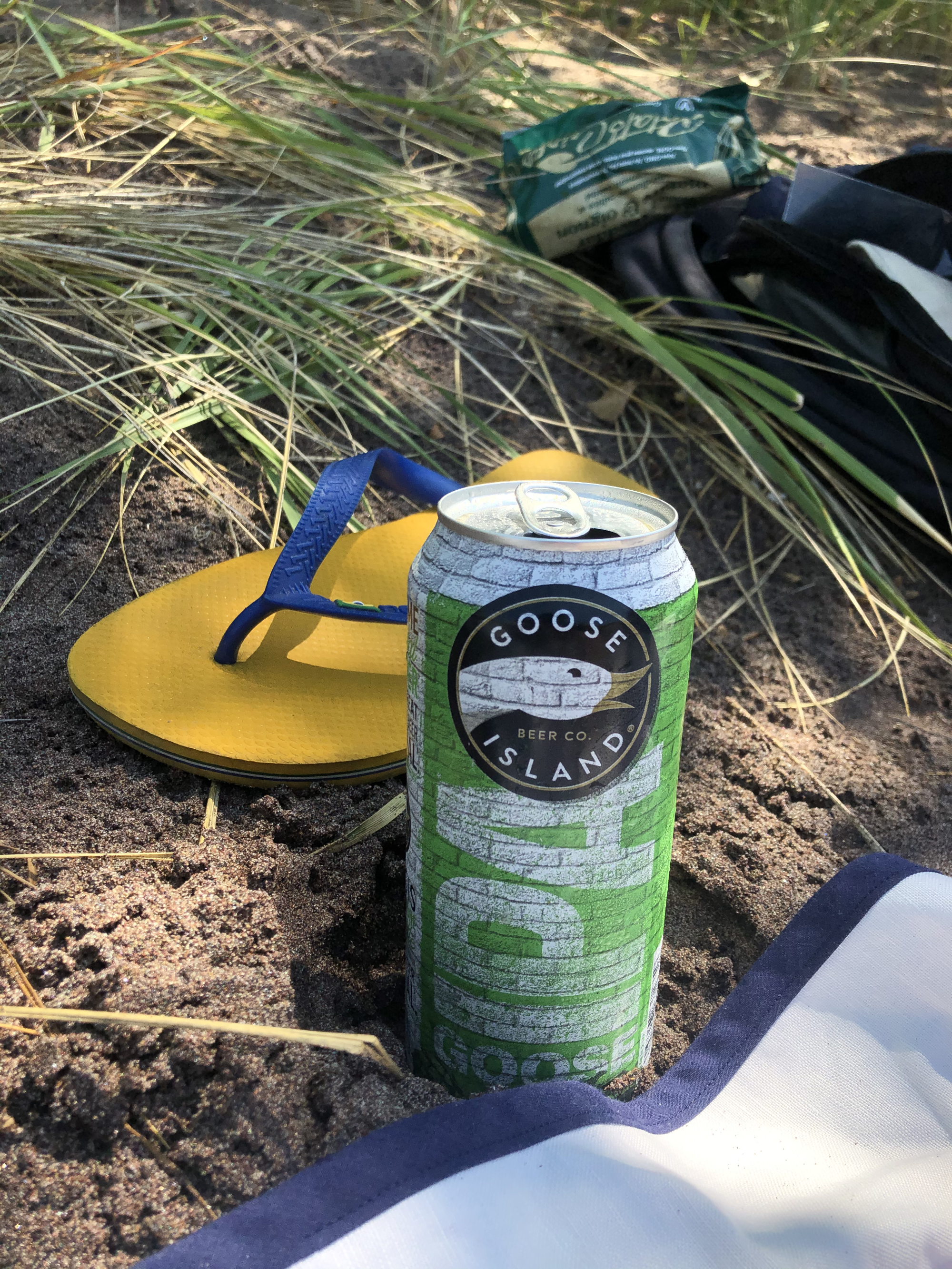 Sur une plage, Canette de bière Goose Island, Sandale Hawaiana jaune, et herbes en arrière-plan.